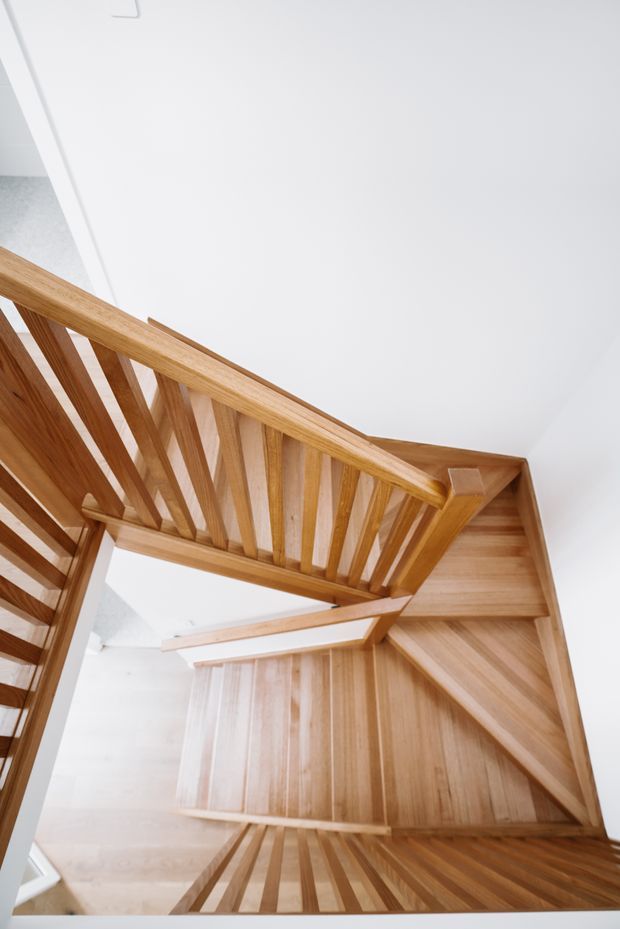A bright, high-angle view looking down through a multi-level timber staircase. The staircase features warm natural wood treads, landings, handrails and closely spaced vertical balusters, set against crisp white walls and ceilings. The geometric zigzag arrangement and open void create a strong architectural focal point.