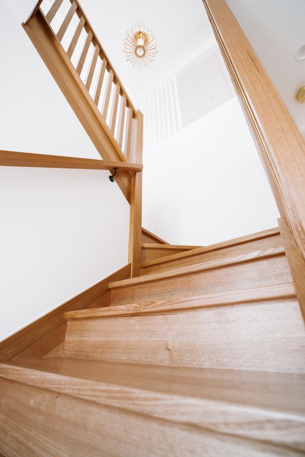 Upward-looking interior photograph of a timber staircase with warm light-wood treads, risers, stringers and balustrades. The stair rises through a bright white stairwell, with a decorative pendant light visible above and strong geometric shadows on the walls.