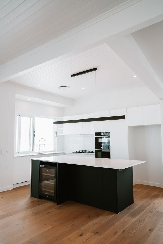 Bright contemporary kitchen with a large black island, white quartz-like benchtop, integrated dark appliances, white vertical wall panelling and cabinetry, pendant strip lighting, and warm timber flooring. The room has a clean, high-contrast, minimalist fit-out with abundant natural light.