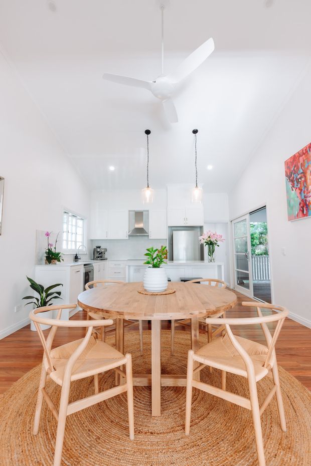 Bright open-plan dining and kitchen interior with a round light timber dining table surrounded by matching woven-seat chairs. The space has white cabinetry and walls, timber flooring, a large natural-fibre round rug, pendant lights, a white ceiling fan, indoor plants, and glass doors opening to an outdoor deck.