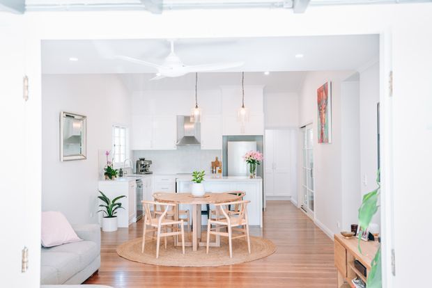 Bright open-plan interior viewed through an open doorway, with a white kitchen, central timber dining table and chairs, light timber flooring, and a compact living area. White cabinetry, pendant lights, ceiling fan, indoor plants, woven rug, and colourful wall art create a fresh, airy residential setting.
