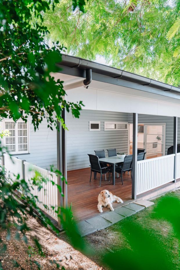 A covered outdoor veranda or patio attached to a light blue weatherboard house, with a timber deck, outdoor dining table and chairs, white balustrades, dark grey posts and roof gutters. The space opens onto a small paved and landscaped garden, with leafy trees framing the view.