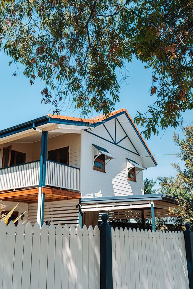 Exterior view of a two-storey white weatherboard home with a prominent gabled roof, decorative dark gable bracing, upper-level balcony, louvred carport, and white picket fencing. The house has contrasting dark blue-green posts, trims, and gutters, with warm terracotta roof tiles and timber balcony decking. Leafy trees frame the sunny residential streetscape.