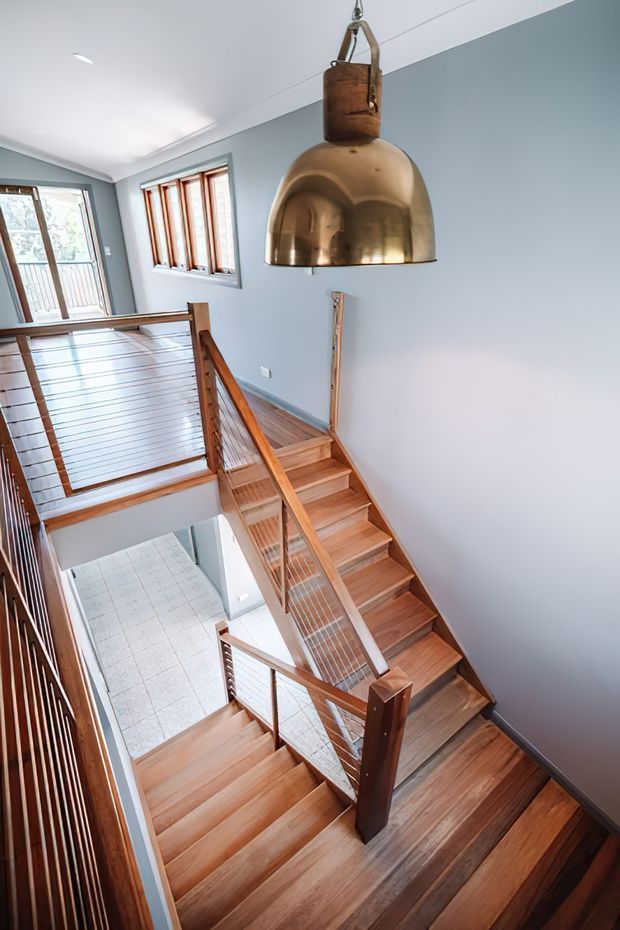 A bright multi-level stairway and landing with warm timber stairs, balustrades, and handrails. The stairwell has pale blue-grey walls, a white ceiling with recessed lighting, timber-framed windows and glazing, and a prominent oversized brass dome pendant light. The open void creates a strong vertical connection between levels.