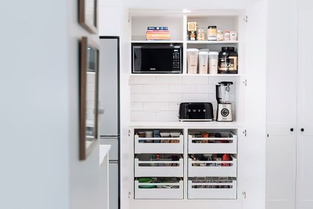 Bright, compact white built-in pantry with open shelves, a microwave, toaster, coffee maker, food containers, and multiple pull-out storage drawers. White subway-tile backsplash and white cabinetry create a clean, highly organised utility space.