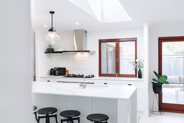 Bright white kitchen with a large island and seating, white shaker-style cabinetry and horizontal wall panelling, a dark benchtop shelf, timber-framed windows and glazed doors, and a prominent skylight. A living area is visible beyond the doorway.