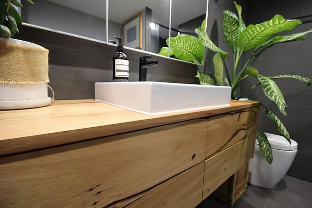 Close-up of a contemporary bathroom vanity with a white vessel basin set into a thick natural timber countertop and matching timber drawers. Dark grey wall tiles, a black tap, mirrored cabinetry, a toilet, and leafy indoor plants complete the scene.