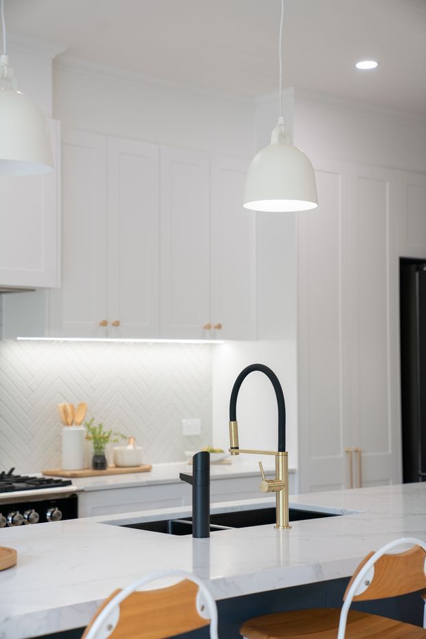 Bright, refined kitchen interior featuring white Shaker-style cabinetry, a herringbone tile splashback, a large white stone island with integrated dark double sink, and contrasting matte black and brass-gold tapware. White dome pendant lights hang above the island, with timber-and-white bar stools in the foreground and a dark range appliance at left.