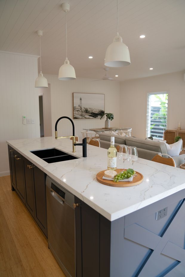 Bright open-plan kitchen viewed from the island, featuring a large white veined stone benchtop, dark navy/charcoal shaker-style cabinetry, integrated sink and dishwasher, brass and matte-black tapware, pendant lights, timber flooring, and a dining and living area beyond. White painted vertical wall panelling and a white painted lined ceiling add a light coastal feel.