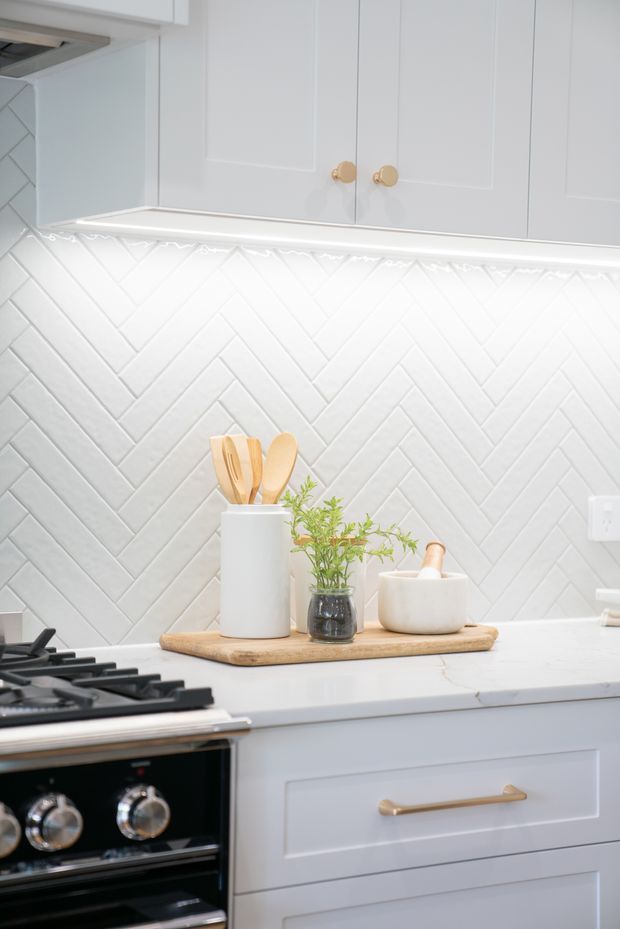 Bright close-up view of a refined kitchen featuring pale blue-grey shaker-style cabinetry, brushed brass knobs and drawer pulls, a white herringbone tile splashback, white benchtop, under-cabinet lighting, and a black freestanding cooktop and oven. A timber tray with utensils and a small plant adds warmth to the otherwise light palette.