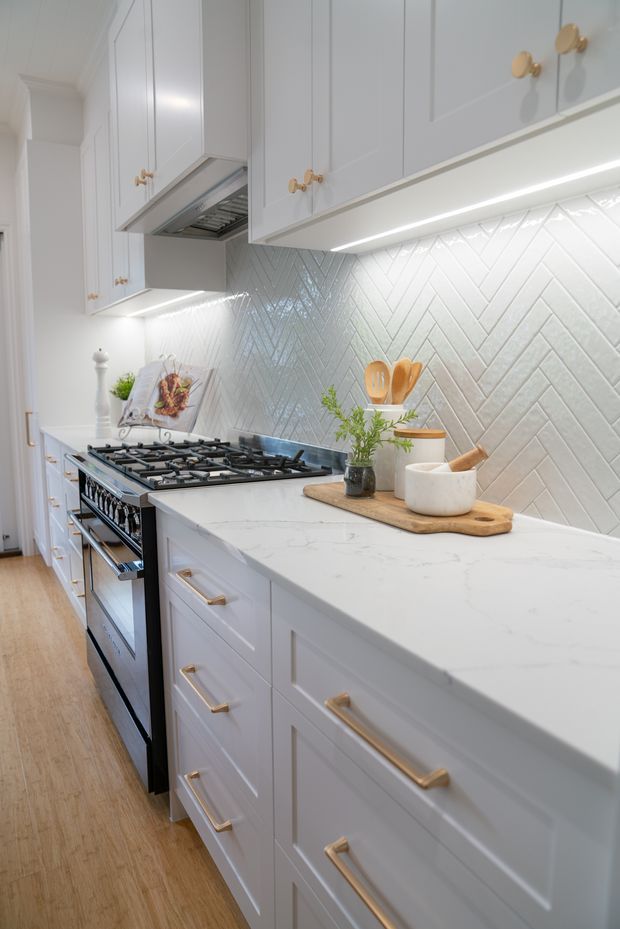 A bright, narrow galley kitchen with white shaker-style cabinetry, brass-gold handles, a large white veined stone benchtop, and a glossy white herringbone tile splashback. A black freestanding gas range sits along the wall, with under-cabinet LED lighting, timber flooring, and a wooden serving board with kitchen accessories on the counter.