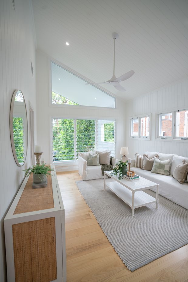 Bright, airy living room with white vertical wall panelling and a white-lined pitched ceiling. Large windows and high-level glazing provide abundant natural light. The room features white slipcovered sofas, a pale timber floor, a large neutral textured rug, a white coffee table, and a slim console with woven-front cabinetry and an oval mirror. A white ceiling fan hangs centrally.