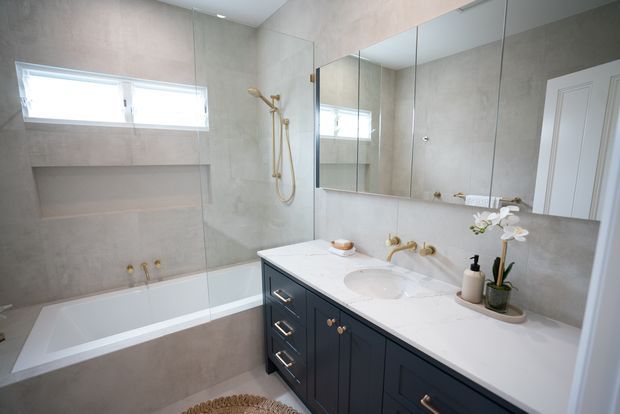 Bright contemporary bathroom with a white built-in bathtub and glass shower screen, large-format warm grey wall and floor finishes, a deep navy-blue double vanity with white benchtop and integrated basin, brass fixtures, mirrored cabinetry, recessed wall niche, and a small orchid arrangement.