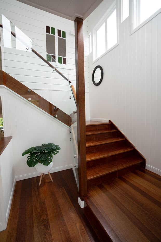 Bright interior stairway with rich dark timber flooring and timber staircase, white horizontal wall panelling, glass balustrades, and a prominent timber post. Tall windows provide abundant natural light, while a decorative stained-glass window and potted plant add visual interest.