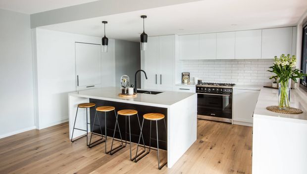 Bright contemporary kitchen with a large white island, integrated white cabinetry, white subway-tile splashback, timber flooring, black pendant lights and four bar stools. The space has a clean, open layout with natural greenery adding a small accent.