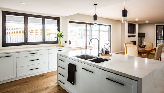 Bright open-plan kitchen with a large white island, integrated double sink, black hardware, pendant lights, timber flooring, dark-framed windows, and white shaker-style cabinetry. A dining area and living space are visible beyond the kitchen.