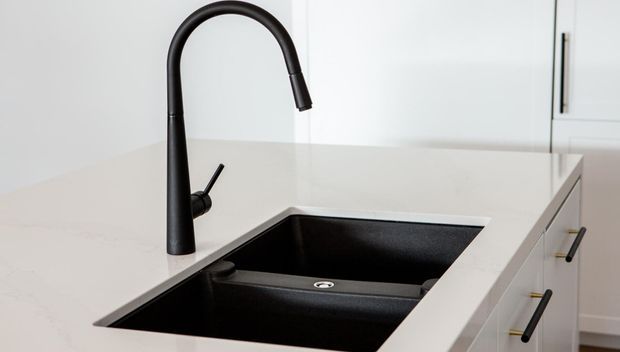 Close-up of a contemporary kitchen work area featuring a large black undermount double-bowl sink and matching tall matte-black gooseneck mixer tap set into a pale stone benchtop. White cabinetry with slim dark hardware is visible in the background.