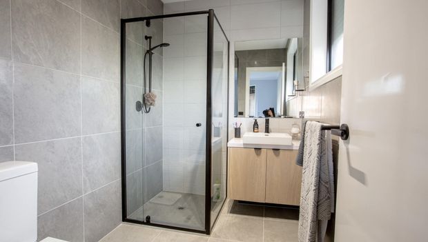 Bright contemporary bathroom with a frameless black-framed glass shower enclosure, large pale grey wall and floor tiles, a floating light-wood vanity with a white vessel basin, and a large mirror. A narrow window provides natural light, while grey towels and minimal fixtures add restrained texture.