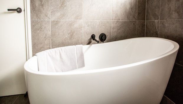 A modern bathroom featuring a freestanding white oval bathtub positioned against large-format grey stone-look wall tiles. Matte black wall-mounted tapware and a simple white door with a black lever handle are visible.