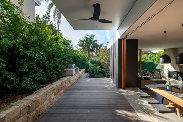 A contemporary tropical residence with a broad timber deck walkway beside dense greenery and a low natural-stone retaining wall. The covered outdoor circulation space opens into a refined dining and living area through large sliding doors, with a ceiling fan, pendant lights, timber dining table, and stone fireplace visible inside. The scene is bright, open, and strongly connected to the landscaped garden.