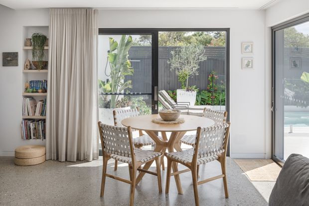 Bright, airy dining area with a round light-wood dining table and woven dining chairs, set beside full-height glazing and a sliding glass door opening toward a landscaped courtyard and pool. Built-in open shelving, full-height curtains, a pale textured floor, and neutral white walls create a relaxed contemporary coastal feel.
