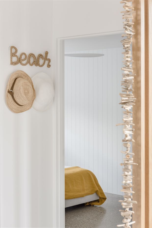 A bright coastal-style interior view through a doorway into a bedroom. The white entry wall features woven rope lettering reading “Beach” and wall-mounted straw hats. A shell garland frames the doorway, while the bedroom beyond has white vertical wall panelling, a mustard ochre bed cover, a pale upholstered bed base, and a grey terrazzo-like floor.