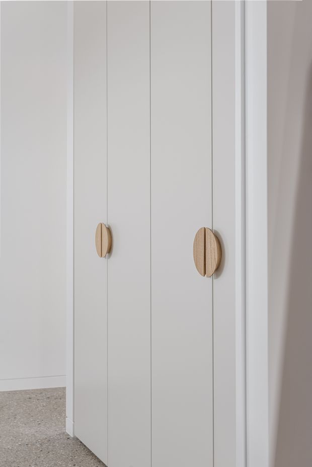 Close-up of a tall white built-in wardrobe or storage cabinet with vertically panelled doors and contrasting light timber oval handles. The cabinet stands against pale walls, with a speckled hard floor visible at the base. The design is simple, clean, and understated.