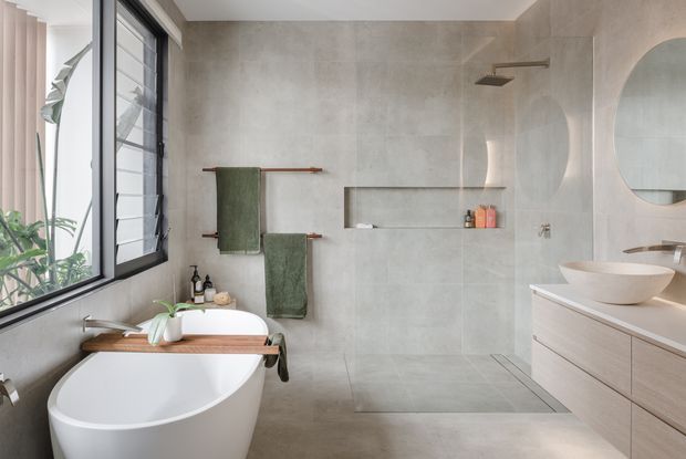 Bright contemporary bathroom with a freestanding white bathtub beside a large black-framed louvred window, a tiled walk-in shower, floating timber-toned vanity with vessel basin, round mirror, recessed shower niche, and green towels. Soft natural light and warm neutral finishes create a calm, refined atmosphere.