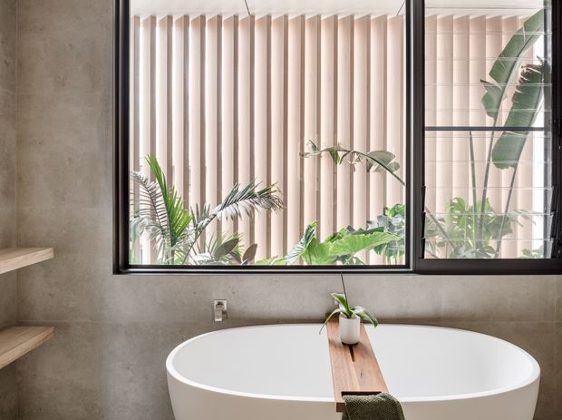 A contemporary bathroom featuring a freestanding white oval bathtub in front of a large black-framed window. The room has warm grey concrete-look wall finishes, timber wall shelves, and a narrow timber bath caddy with a small potted plant. Vertical privacy fins and lush tropical foliage are visible beyond the glazing, creating a calm, natural atmosphere.