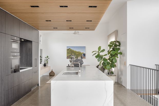 Bright contemporary kitchen with a large white stone island, integrated stainless-steel sink and tap, dark timber-look cabinetry with built-in appliances, and a slatted timber ceiling with recessed lighting. The kitchen opens toward a stair landing, with white walls, terrazzo-like flooring, indoor plants, and artwork adding warmth.