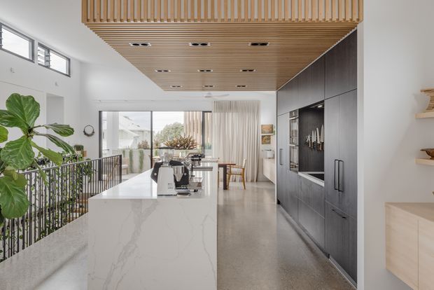 Bright contemporary kitchen with a large white stone island, integrated black appliances and dark full-height cabinetry. A timber slatted ceiling feature adds warmth above the open-plan space, with polished concrete-style flooring, white walls, indoor planting, and glazing opening toward an outdoor area.