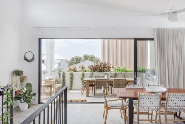 Bright contemporary open-plan dining and living area opening onto a landscaped outdoor terrace. The room features a large glazed sliding opening, pale polished concrete flooring, white walls, indoor plants, a timber dining table with woven chairs, and a ceiling fan. A covered outdoor dining area and planter-lined courtyard are visible beyond.