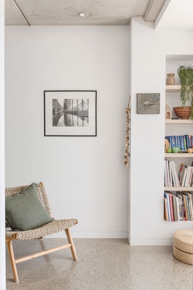 Bright minimalist interior corner featuring a woven timber-framed lounge chair with a muted sage cushion, a framed monochrome artwork, built-in open shelving filled with books and decorative objects, and a woven round ottoman. The space has white plaster walls, a pale exposed concrete ceiling, and a polished speckled concrete floor.