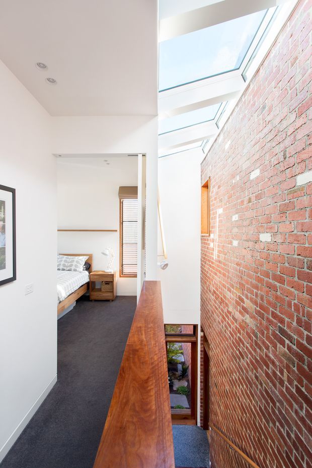 A bright, narrow upper-level hallway or landing beside a staircase, featuring a long polished timber balustrade, exposed red brick wall, white plaster walls, dark carpet, and large overhead skylights. A bedroom is visible through the open doorway at the end, with a timber bed frame and bedside table. The space has strong natural light and a contemporary renovation feel with retained masonry.