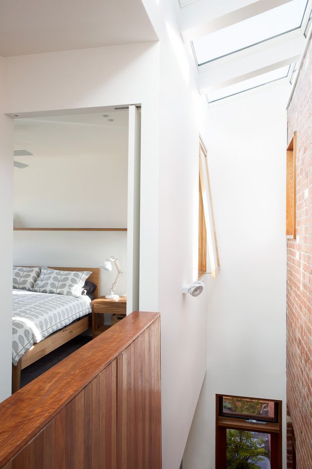 Bright, narrow multi-level interior void or stair landing with a bedroom visible through a wide opening on the left. The space features a tall white-painted wall, a red exposed-brick wall on the right, a series of high clerestory/skylight windows, timber-framed windows, and a substantial dark timber balustrade or built-in guardrail overlooking the lower level. The bedroom has timber joinery, patterned bedding, and a small bedside table.