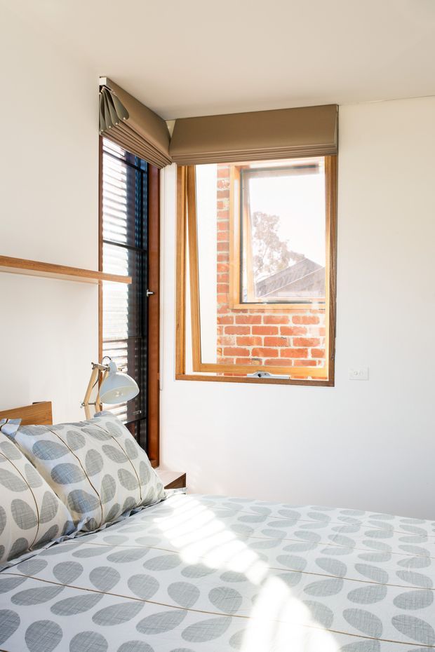 Bright compact bedroom with a bed dressed in a pale grey and white leaf-patterned cover, a simple bedside lamp, white walls, timber-framed windows, and roller blinds. Exposed red brickwork is visible outside the window, with strong natural sunlight entering the room.