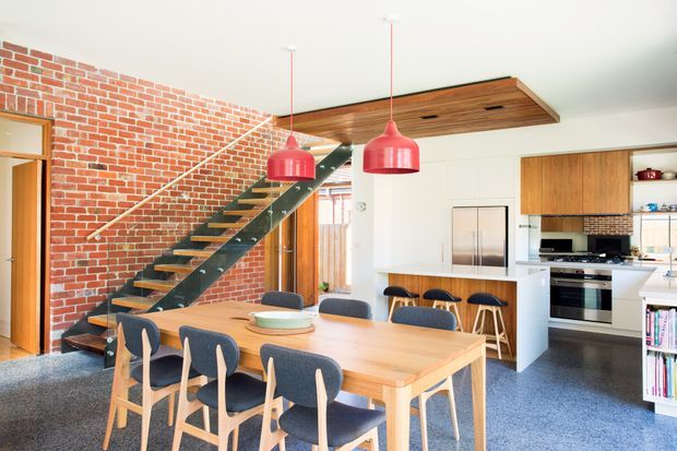 Open-plan dining and kitchen interior with a large timber dining table and upholstered timber chairs in the foreground. A dramatic black metal and timber staircase runs alongside an exposed red-brick wall, while the contemporary white kitchen includes timber cabinetry, stainless-steel appliances, a peninsula with stools, and a timber-lined ceiling feature with pendant lighting.