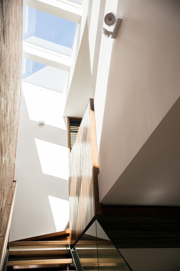 A bright, narrow stairwell viewed upward, featuring a timber staircase, dark timber accents, a clear glass balustrade, tall white walls, and large overhead skylights casting strong geometric shadows. A sculptural timber screen or partition rises alongside the stairs, adding warmth and texture to the otherwise crisp, light-filled space.