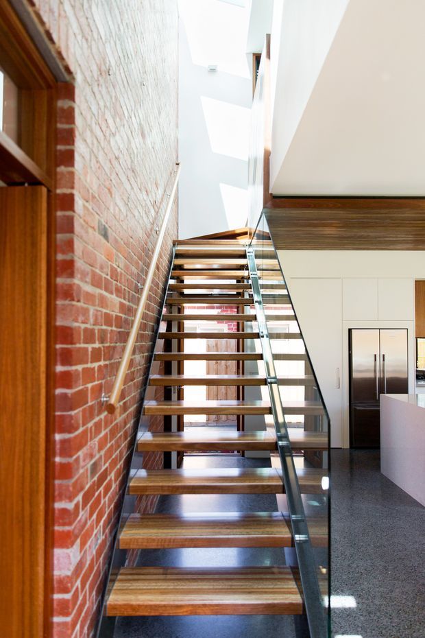 A bright, narrow stairway with open-riser timber stairs, a clear glass balustrade, and a slim dark handrail. The stair runs alongside an exposed red brick wall and leads toward an upper-level landing. A contemporary kitchen with white cabinetry, timber trim, and a stainless-steel refrigerator is visible to the right. Skylights or clerestory glazing create strong natural illumination.