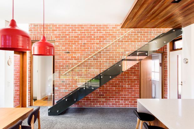 Bright contemporary interior featuring a dramatic black steel and glass staircase against an exposed red brick wall. The space combines a timber-lined ceiling, pale walls, grey carpet, white cabinetry or benchtop, and vivid red pendant lights, with a dining area visible at left and a kitchen or work counter at right.