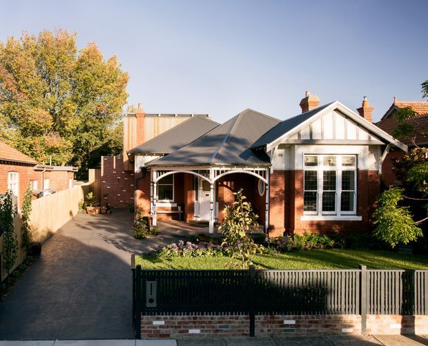Street-facing view of a single-storey brick residence with a front gabled porch, decorative timber gable detailing, corrugated metal roof sections, tall divided-pane windows, brick chimneys, a landscaped front garden, and a driveway leading beside the house. The home combines traditional detailing with a compact urban suburban form.