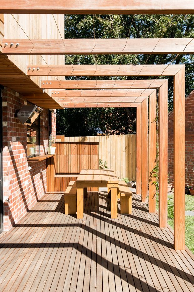 A sunny outdoor timber deck courtyard beside a red brick wall, furnished with a built-in timber dining table and benches. Repeated timber overhead beams create a pergola-like covered walkway, with timber screening and fencing at the rear, potted plants, and strong linear shadows across the decking.