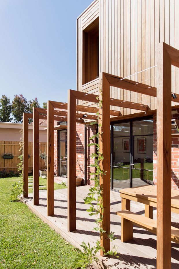 Sunlit exterior view of a contemporary two-storey brick house with warm timber vertical cladding and a timber-framed pergola extending over a raised outdoor deck. The pergola includes climbing plants, integrated lighting and a timber picnic-style table, while large glazed doors connect the deck to the interior. A fenced garden is visible alongside the building.