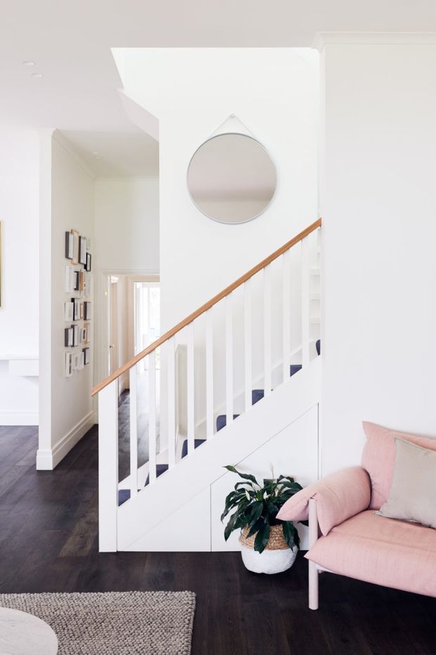 Bright white residential hallway and stairway with white-painted balustrades, a warm timber handrail, dark timber flooring, built-in under-stair storage, framed wall display, and a round wall mirror. A blush pink armchair and potted plant add soft colour in the foreground.