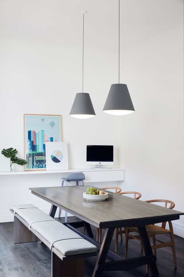 Bright contemporary dining and work area with a long dark timber dining table, upholstered bench seating, wooden chairs, and two charcoal pendant lights. A minimalist white built-in desk with computer sits against the wall, accompanied by colourful framed artwork and a small plant. The room has white walls, dark timber flooring, and a clean, airy composition.
