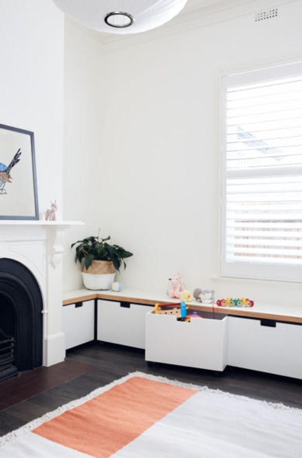 Bright, compact children's room with white walls, a black traditional fireplace, built-in low white storage and desk cabinetry beneath a timber-toned countertop, a leafy plant, toys, framed artwork, plantation shutters, and a soft peach-and-white rug. The room has a clean, playful, lightly traditional interior.