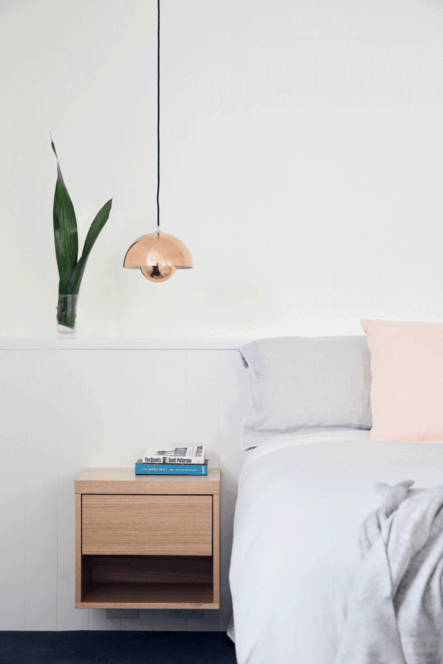 Bright, minimalist bedroom vignette featuring a white bed with pale bedding and a blush cushion, a wall-mounted timber bedside table, a copper pendant light, and a tall green plant on a white built-in ledge. The room has clean white wall panelling and a calm, softly styled aesthetic.