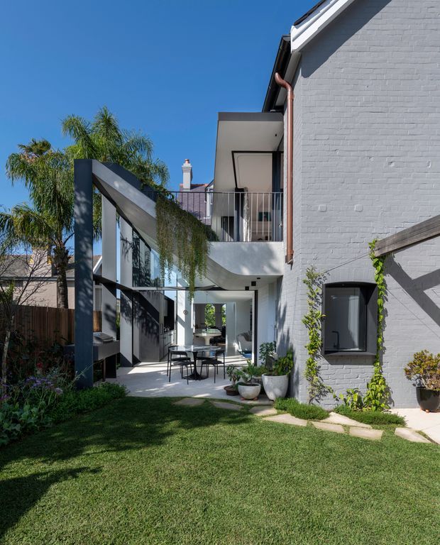 A sunny courtyard garden beside a renovated white-painted brick building. The view highlights a two-level outdoor living area with a covered patio, glass walls and doors, balcony, dark metal-framed glazing, climbing greenery, potted plants, lawn, and a contemporary extension linked to the existing structure.