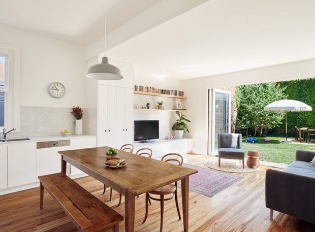 Bright open-plan kitchen and dining area leading into a living room and garden. The space features white cabinetry, a pale stone backsplash, warm timber flooring, a rustic timber dining table with matching bench, bentwood chairs, open shelving, and a large glazed opening to the outdoor area. Neutral finishes and abundant natural light create a relaxed, inviting atmosphere.