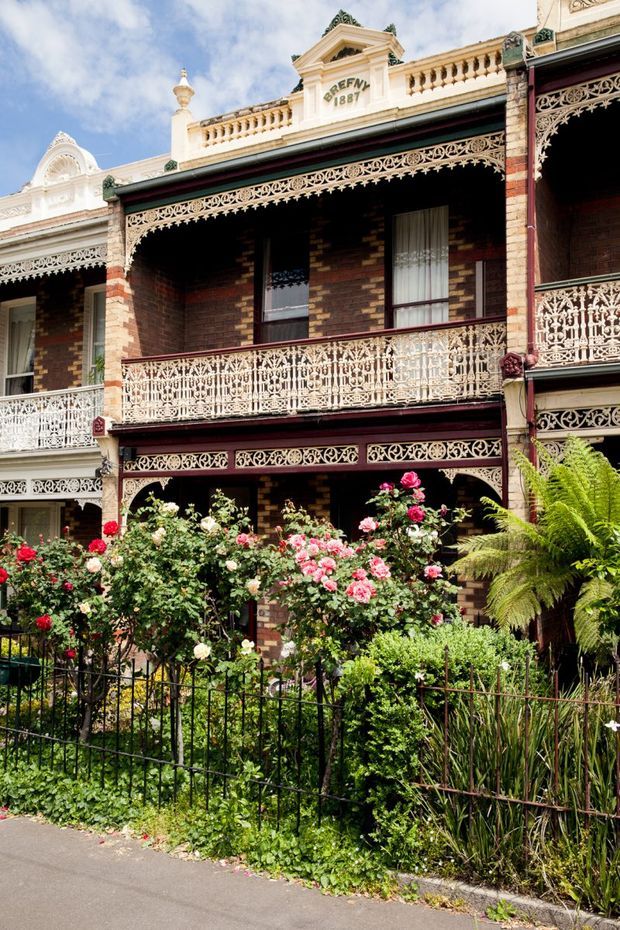 Street-facing historic terrace house with dark red and brown brickwork, ornate cream-painted cast-iron lacework across the balcony and verandah, decorative parapets and balustrades, and a lush cottage-style front garden filled with roses and ferns. The facade has strong Victorian-era detailing and a clearly visible date plaque reading 1887.